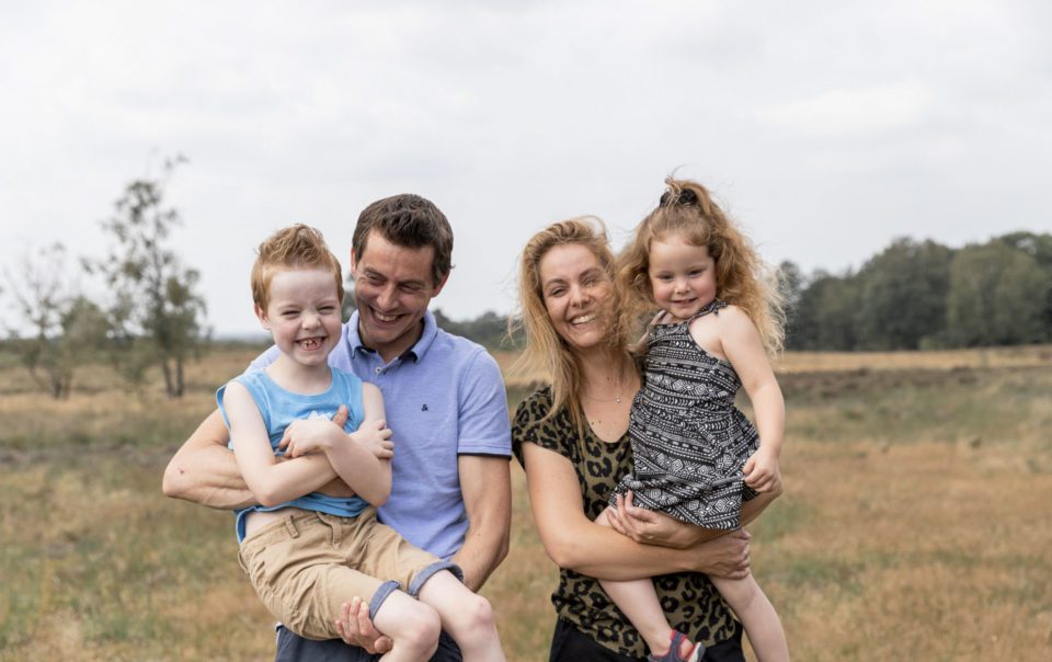 Een stralende familie van vier geniet van een ontspannen moment in het park van Radio Kootwijk, omringd door groen, terwijl ze samen oprecht lachen voor de camera tijdens hun familie fotoshoot.
